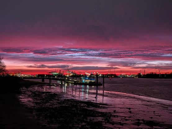 Sonnenaufgang mit kräftigen roten Farben, purpurroten Wolken. Die Lichter vom Fähranleger schimmern vor dem imposanten Himmel. Die Farben werden vom nassen Sandstrand teilweise gespiegelt.