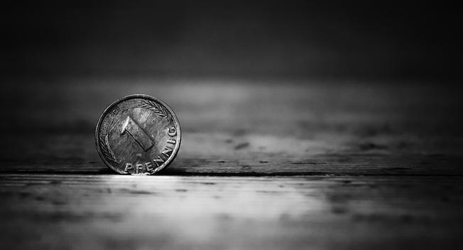 Man sieht eine 1 Pfennig-Münze die senkrecht wegen einer kleinen Spalte im Holzboden steht. Sie ist klar und deutlich zu sehen während der Hintergrund wie auch der Vordergrund unscharf sind. Das Foto ist in Schwarzweiß.