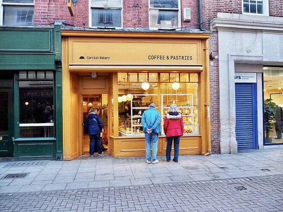 a Cornish bakery shop with wooden exterior and “coffee and pastries” on the sign. Two people are standing at the window looking through into the shop while someone else wanders in through the door.
