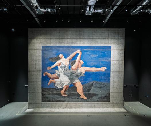 A large theatrical backdrop painting displayed in a darkened gallery space, showing two figures running or dancing against a vibrant blue sky. The neoclassical-style figures wear flowing white and gray drapery, depicted in earth-toned flesh colors. They move dynamically across a dark landscape with mountains visible in the background. The artwork is painted on a textured, cream-colored canvas with visible wear and aging, mounted on a concrete or stone-like wall and dramatically lit from above by gallery spotlights.