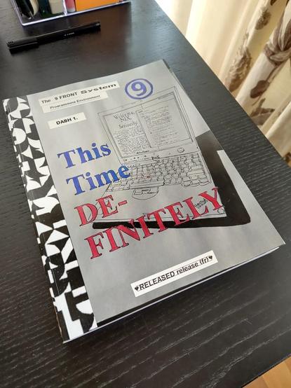 black table with a book on top. It says "The 9 FRONT System Programming Environment" DASH 1. "This Time DE-FINITEY," featuring a hand-drawn black and white thinkpad. The book's cover has bold text in blue, red, and black, with a white and black geometric pattern on the left. At the bottom it says "RELEASED release (fr)" Some of the titles have been created with a Dymo sticker maker and the original title text was filled with a blue and red marker.