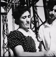 Araceli Sánchez Urquijo in B&W photo, stood next to young man pointing something out to him on a plan. Complicated very large industrial metal structure in the background, wearing dark short sleeved dress with white polka dots and a white collar and frill. Young white woman with dark hair drawn back into bun