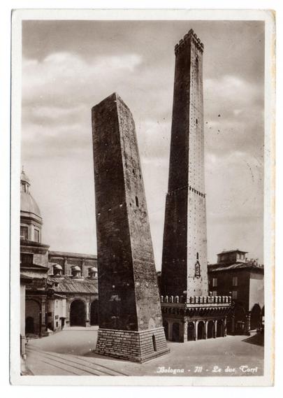The black and white photograph features two prominent, ancient stone towers standing in a town square. The tower on the left is shorter with rectangular sides, while the one on the right is taller and more slender, topped by what appears to be remnants of battlements or fortifications suggestive of medieval architecture.

In front of these structures lies an open plaza that leads towards another building featuring classical domed roofs in the background, hinting at a church or public edifice. The ground shows signs of wear with visible stains and patches indicating age and use over time. An archway can be seen leading into what might have been additional buildings or courtyards.

Text overlay on the image states "Bologna - II- Le due Torri," which translates to Bologna - Parts II, The Two Towers in English, identifying these towers as significant landmarks within that city's historical context.