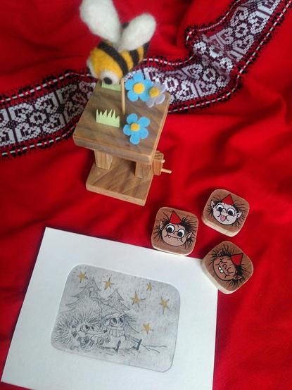 Photo présentant des objets et tirages sur tissu rouge. En haut, une sculpture-abeille qui gigote au-dessus des fleurs. À droite des magnets avec des têtes de lutins qui font des grimaces. En bas, un tirage de gravure tetrapak qui représente un lutin et un hérisson regardant les étoiles. Les étoiles sont peintes en doré.