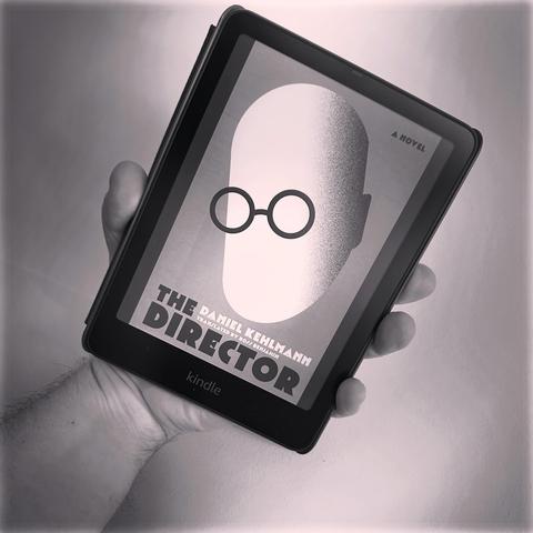 A hand holds a Kindle displaying the cover of the novel "The Director" by Daniel Kehlmann. The cover features a minimalist design with a faceless figure and round glasses, predominantly in black and white.