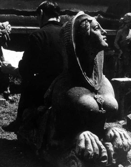 The black and white photograph depicts a person sitting on the ground beside an imposing statue. The figure appears contemplative, wearing dark clothing that contrasts with their pale complexion due to the monochrome effect of the photo.

This individual is seated next to what seems like part of another sculpture or perhaps even another person in close proximity, as suggested by partial outlines and shadows visible around them.

The focus of the image is on a large statue portraying an Egyptian woman. The figure exudes an aura of antiquity with detailed carvings adorning its body; it's dressed traditionally, featuring what appears to be a headdress or headpiece indicative of ancient Egyptian royalty or deities.

This sculpture showcases intricate craftsmanship: there are visible folds in the garment that could represent cloth draped over the shoulders and torso. The statue is seated on ornate stone-like bases for both feet, adding to its grandeur.

The background features darker tones which might be part of an outdoor setting with other sculptures or structures faintly discernible but not clearly defined due to the depth of field used in capturing this image.

There's a visible signature mark at the bottom right corner, indicative of it being taken by a professional photographer. This particular photograph is associated with Donzelli Pietro’s work and can be referenced for further context via the provided URL link.