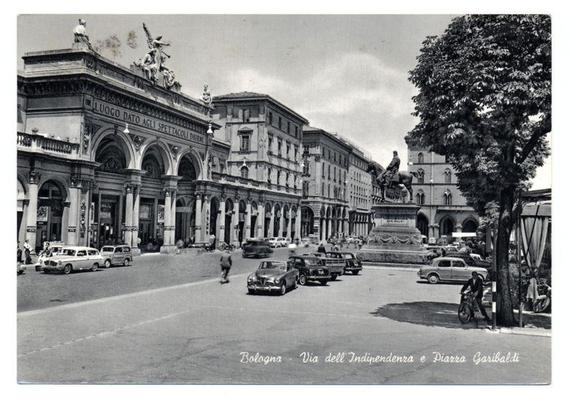 The image is a black and white photograph depicting an urban scene with historical architecture. Prominent in the center is a large building adorned with arches, columns, and statues on its facade. Above this structure, there's an inscription "LUOGO DATO AGLI SPETTACOLI," which translates to "Given as for Spectacles." To the right of the central building stands another edifice featuring classical architectural details such as a pediment with sculptural elements.

In front of these buildings is a busy street bustling with vintage cars and pedestrians. Several vehicles, including sedans and an SUV, are parked or in motion along the road. Pedestrians can be seen walking on the sidewalks or crossing at intersections.

A notable feature includes a statue atop a pedestal situated near the center-right part of the image; this figure appears to represent a historical individual mounted on horseback, with accompanying inscriptions that read "C.A.BALD" and another inscription too small for clear reading. The surrounding buildings are consistent in style, featuring similar ornate details.

Trees line parts of the street scene, adding natural elements amidst the urban environment. In the foreground, to the right side, a cyclist is captured mid-motion on the road, with one car following closely behind him. Other pedestrians can be seen engaging various activities along the sidewalk or crossing paths in crosswalks.

The photograph car [...]