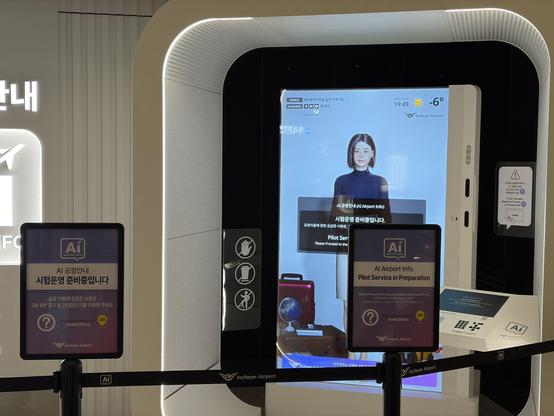 An AI information kiosk at Incheon Airport, featuring a digital avatar providing assistance. Two signs indicate that pilot services are in preparation and provide directions for usage. The display shows current weather, time, and language options.