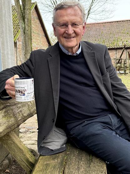 Mann mit Brille liest Zeitung auf einer Holzbank vor einem Backsteinhaus.
