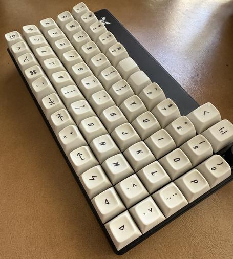 An ortholinear mechanical keyboard with a black case and white sculpted keycaps with black legends at a low 60 degree angle so the sculpted keycap angles are more obvious. The layout of 5 main rows of 1U keys by 12 keys across is augmented by two larger keys and a volume knob above
the top right of the main keyboard. The layout pushes the printable characters to the edges of the keyboard and uses almost only the lowest row for modifiers and keys for enter, escape, tab, backspace and cursor
movement. Notably two keys on the bottom
row each one key away from the center columns use a high sculpted profile from the top row and are expected to be where the thumbs rest. They are the dash/underscore on the left and the equals/plus sign keys on the right. The center column is all punctuation or symbol keys for the first four rows, except the top row where the middle columns have unlabeled keys. The layout of letters is the same as US English layouts with a Shift key left of Z on the fourth row down, and P in the right column on the second row down. There are cursor
down and cursor right keys in the rightmost columns of the bottom row, with a function key to the left of them. The keyboard is sitting on a leatherette semi-reflective material with sunlight glare behind it.