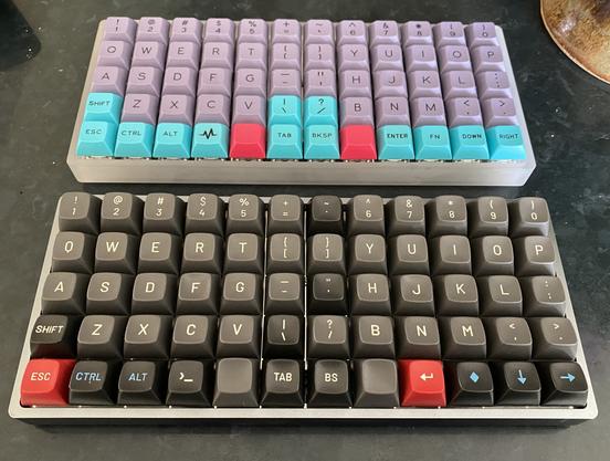 Two more simple 12x5 ortholinear keyboard original Bucktooth layout examples, one with purple flat keycaps for most printable characters and cyan keycaps for modifiers and function keys, and another with mostly grey sculpted keycaps for printed characters but some are black including the backtick in the top row and apostrophe in the middle and the backslash in the lower center. Two keys are red, the ESC key on the lower left and the enter symbol key on the right side next to the right space key.