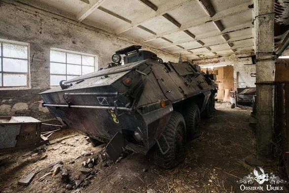 Inside an old, dilapidated building sits a large, black military vehicle. Windows let in natural light, illuminating the dirt floor and the