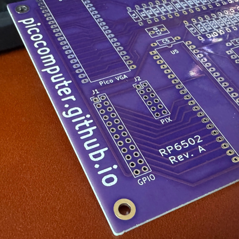 A close-up photograph of a purple printed circuit board labelled “picocomputer.github.io”. The board has white silkscreen text marking headers such as “Pico VGA”, “PIX”, and “GPIO”, along with the designation “RP6502 Rev. A”. The PCB has unpopulated through-hole pads and a mounting hole at the corner. The surface traces and via patterns are clearly visible.