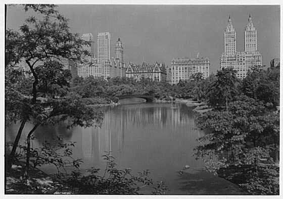 The image depicts a black and white photograph of Central Park, showcasing the iconic skyline reflected on the tranquil waters. The scene captures tall skyscrapers like San Remo and Lexington Towers in Manhattan across the lake's reflective surface. In the foreground are trees with branches reaching into the water, adding texture to the composition. A bridge spans over part of the park's pond, connecting two green spaces amidst urban buildings. This historical photo is credited to Samuel H. Gottsch from June 4th, 1932, as detailed in New York city views collection.