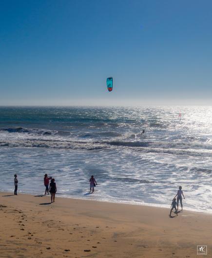Color photo of a group of people standing on a beach at the water’s edge watching a kite surfer amongst the waves offshore on a sunny day under clear blue skies.