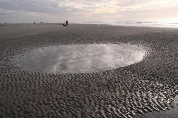 Wijdse blik op een oneindig groot geribbeld strand. In een rond poeltje van achtergebleven zeewater wordt de bewolkte lucht weerspiegeld. 
In de verte een persoon met een hond, zij staat op het punt een bal weg te gooien met een slingerstok.
Daarachter de Noorzee.
Heel in de verte langs de vloedlijn kleine silhouetten van wandelende mensen.