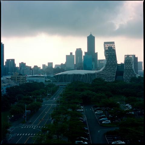A cityscape under a cloudy sky with modern skyscrapers and a distinctive architectural structure in the foreground. A road with traffic lights and parked cars runs through a tree lined area. The scene suggests an urban environment during dusk.
