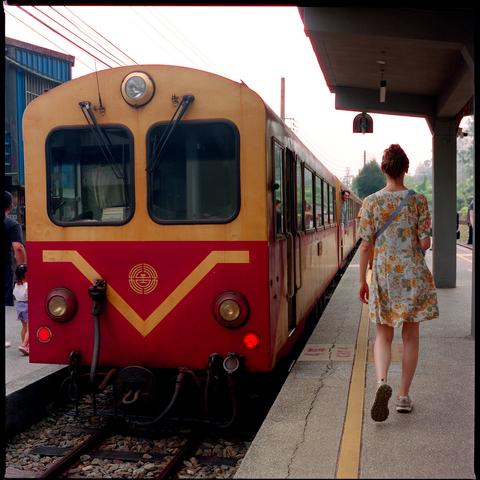 A woman in a floral dress walks along a train station platform toward a red and yellow train. The train has a V shaped logo on its front, and people are visible near the platform and inside the train. Overhead wires and a covered platform structure are also visible.