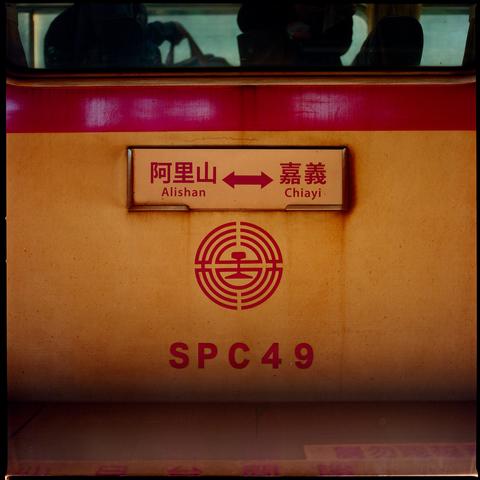 A close up of a Taiwan Railway train car, showing a route sign indicating travel between Alishan and Chiayi. Below the sign is the Taiwan Railway Administration logo and the car number SPC49. The train's body is orange with a red stripe. Reflections of passengers are visible in the window above.