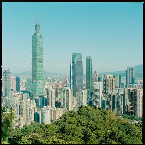 A panoramic view of Taipei's skyline featuring the iconic Taipei 101 tower rising above a dense cluster of modern skyscrapers and residential buildings. Lush green trees frame the foreground, while distant mountains are visible under a clear blue sky.
