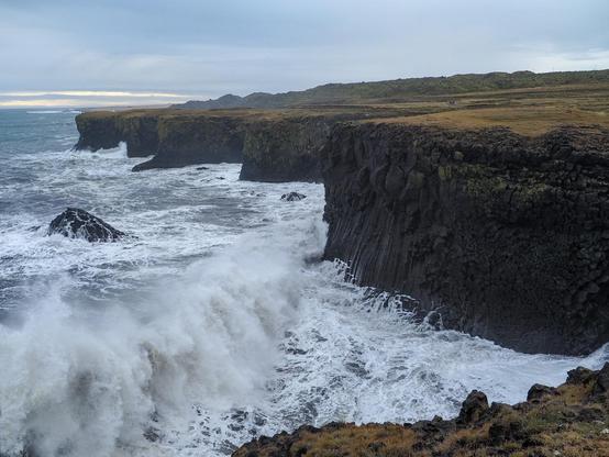A big wave rolling towards black sea cliffs on a stormy, cloudy day.