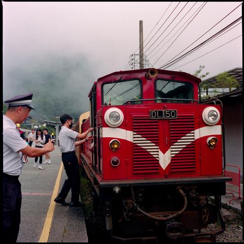 Red diesel locomotive DL 50 at station platform. Crew member assists passenger boarding. Misty mountain backdrop. Overhead power lines visible. Classic train design with white chevron marking.