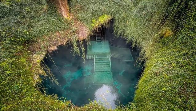 Aerial view of Devil's Den spring in Florida showing crystal-clear turquoise water inside a circular sinkhole surrounded by dense green vegetation, with wooden stairs and platforms descending into the water.