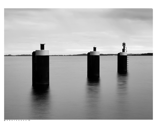 Aufnahme in schwarzweiß, drei massive Zylinder ragen aus dem Wasser des Hafenbeckens heraus, Anlegemöglichkeiten für Schiffe, durch Langzeitbelichtung ist die sonst wellenreiche Oberfläche des Wassers glatt und verschwommen, hellgrauer Himmel darüber
