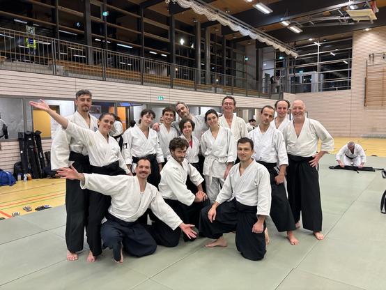 14 pratiquant·es d'aïkido posent pour la caméra avec des visages souriants, sur un tatami à l'intérieur d'un grand gymnase.
---
14 aikido practitioners are doing a group pose on a tatami, inside a large sports hall.