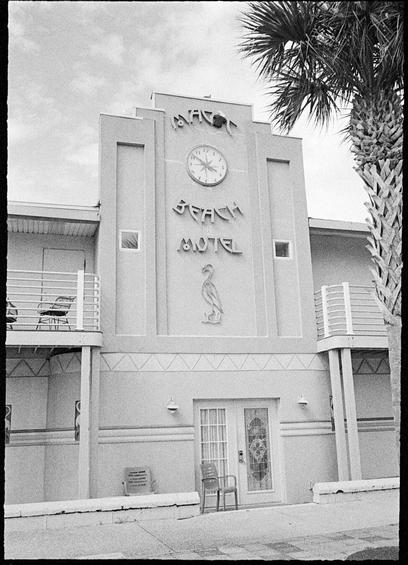 A cement building in an art deco style with a towering front and a sign that says “Magic Beach Motel” where the i in Magic is a rabbit coming out of a hat. There is a flamingo under the word “Motel”, and a clock with a starburst at its center between “Magic” and “Beach”. Two cheap plastic chairs sit in front of a double door under the sign. Bits of flamingoes in relief on the walls are visible.