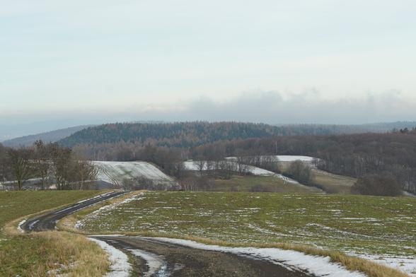 Hügel mit kaum-befestigter Straße, die sich durch die Landschaft windet. Im Hintergrund Wälder und Wolken