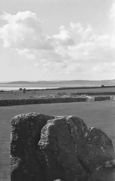 View over some flat, upright rocks, some fields divided by dry stone walls, a bag in the sea, and some hills behind that bay. There's also sheep. And fluffy clouds.