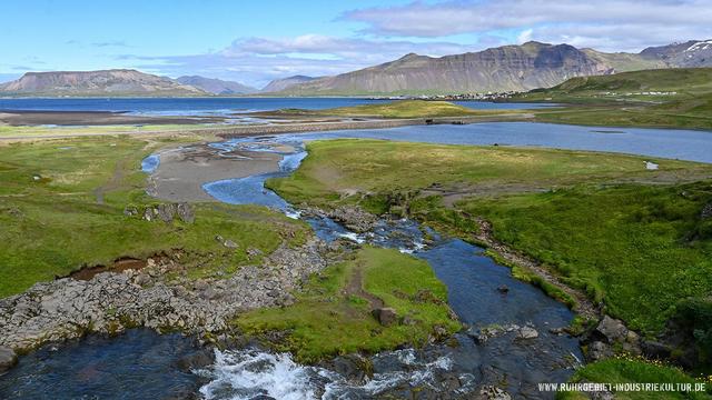 Weitläufige isländische Fjordlandschaft mit einem blauen Fluss, der durch grüne Wiesen fließt, im Hintergrund dramatische Berge und eine kleine Siedlung am Ufer