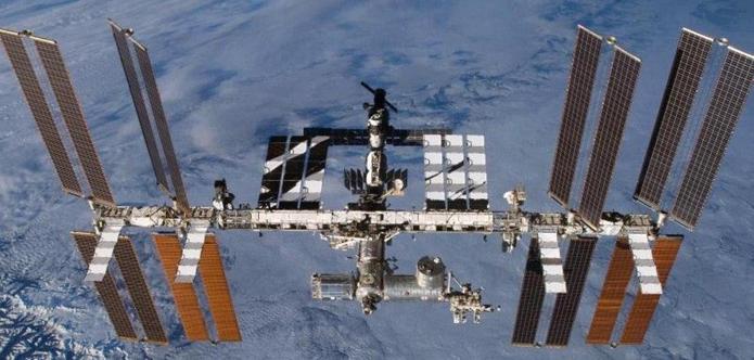 Bild der Internationalen Raumstation (ISS), umgeben von der Erdoberfläche. Die Station zeigt mehrere Solarpanel-Arrays an den Seiten und verschiedene Module in der Mitte. Der Himmel erstreckt sich in einem weiten Bereich um die ISS.