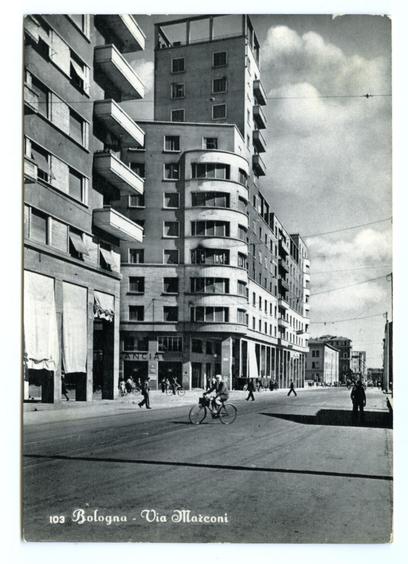 This black and white photograph depicts a street scene with mid-20th-century architecture. Several multi-story buildings line the road, showcasing various architectural styles from Art Deco to modernist influences. The building on the left has an angular design with balconies jutting out at different levels, while the one in the center features curved edges and large windows that reflect sunlight.

On the street, a cyclist rides past two individuals who appear to be walking; their motion is captured as blurred lines due to the long exposure of the photograph. In the background, more pedestrians are seen strolling or standing on the sidewalk near storefronts with signage, including one labeled "ANCIA." The sky above has scattered clouds.

In the foreground at the bottom right corner, there's a handwritten caption that reads "103 Bologna - Via Maconni," indicating the location and possibly an address. The overall composition of this image suggests it was taken in a European city during or shortly after World War II, judging by the architectural styles and urban landscape presented.

The photograph has some signs of wear on its edges, suggesting that it might be from an older archive collection.