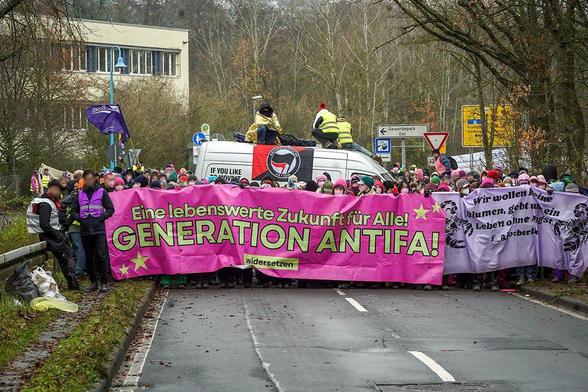 Ein Teil der Demo bei einer Blockade mit dem pinken Transparent des "Widersetzen" Bündnisses mit dem Text "Eine lebenswerte Zukunft für Alle - Genaration Antifa". Daneben ein weiteres Transparent mit dem Text "Wir wollen keine Blumen, gebt uns ein Leben ohne Angst"