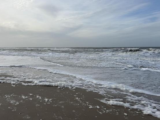 Eine Küstenszene mit Wellen, die gegen den Sandstrand schlagen und unter einem bewölkten Himmel Schaumkronen und brechende Wellen erzeugen.