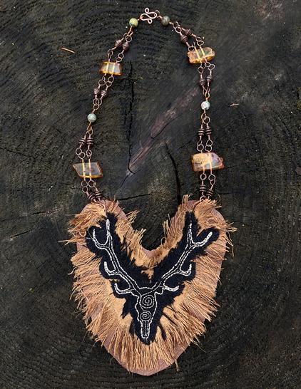 A necklace featuring a large hand embroidered stag on silk and leather, hanging from copper wire adorned with amber, copper, and green jasper beads.