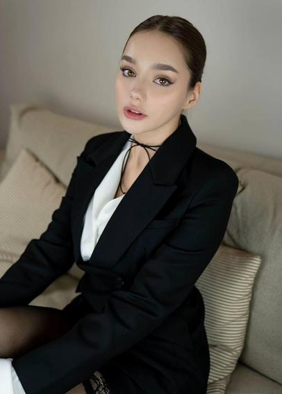 A woman sitting on a couch wearing a black blazer over a white top, with her hair tied back. She looks at the camera with a calm expression. Lace stockings are partially visible, adding a subtle suggestive element.
