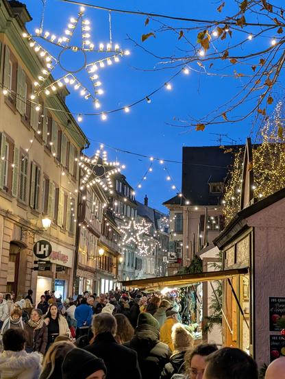 a busy narrow street with white courtyard Christmas star lights hanging above, and market stalls up the right.