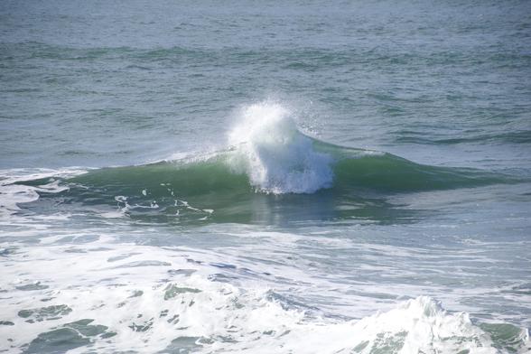 View of a small wave in blue-green ocean water, with somewhat smooth but still choppy water behind it and white foamy small swells in front of it.

There is a small bulbous explosion of white water in the middle of the wave, causing a rounded top with splashing droplets flying up from it  and a white roiling cascade in front of that. All of that is springing from the center of what appears to be a small simple curling wave. 
(It’s actually two similar waves coming in from left and right and crashing together in the middle.)