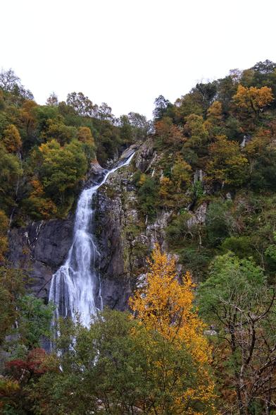 A breathtaking view of a cascading waterfall nestled in a forested mountain landscape. The waterfall descends in multiple tiers, surrounded by lush greenery and vibrant autumn foliage in shades of orange, yellow, and green. The rocky cliff face contrasts with the flowing water, creating a serene and picturesque natural scene. The sky above is overcast, adding a soft, diffused light to the tranquil atmosphere.