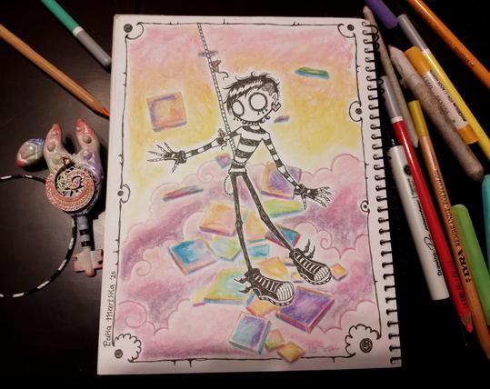 Foto de un cuaderno abierto en el que se ve una ilustración de  una caricatura emo en blanco y negro, de una persona atada por el estómago siendo levantada en el aire entre nubes rosadas. Alrededor del personaje hay baldosas de colores sueltas que parecen estar flotando. A ambos lados del cuaderno hay lápices de colores y pasteles, y también hay una llave de fantasía de los mismos colores.