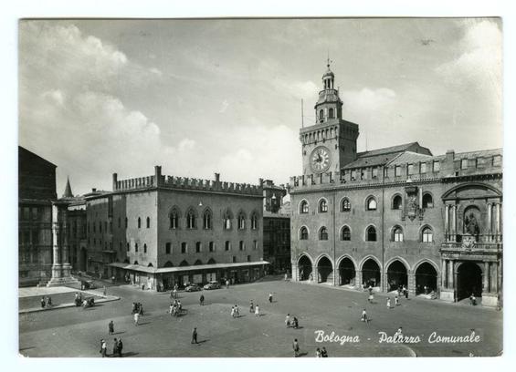 A black and white photograph of a bustling urban square in Bologna, Italy. The image features historic buildings including the Palazzo Comunale with its clock tower prominently displayed at the center-right side. People are scattered throughout the open plaza engaging in various activities such as walking or standing around.

The text "Bologna - Palazzo Comunale" is written across the bottom right corner of the photo, suggesting that this image captures a significant historical and architectural landmark within Bologna's cityscape.