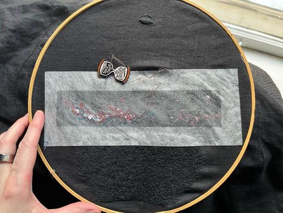 Lia’s fingers show a wooden embroidery hoop with black linen with a rectangle of dissolvable material on which is printed the Milky Way in black and white with some red and blue markings. Some tiny single strand seed stitch and French knots in white, peach, oranges, and dark browns and grays have been stitched. Below the Milky Way is black on black whipped backstitch that looks a bit like flames, it’s pubic hair. An hourglass-shaped needle minder that reads “this is taking forever” is attached to the cloth.