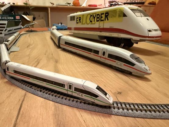The image features several model trains on a wooden surface, including three ICE trains in varying sizes. The tracks are arranged in a curved layout, and there are boxes and other items in the background.