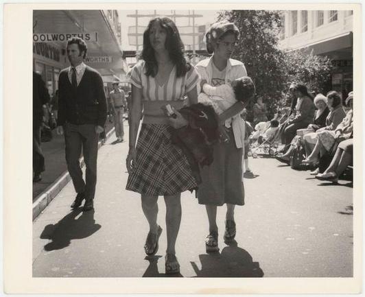 A black and white photo capturing a bustling street scene with multiple people. Two women are prominently featured in the center; one wearing a striped top, plaid skirt, and sandals is walking alongside another woman holding a child dressed in dark clothing. The background shows several individuals seated on benches or standing, suggesting this may be a public gathering area such as a mall's outdoor seating section. A Woolworths store sign indicates commercial activity nearby.