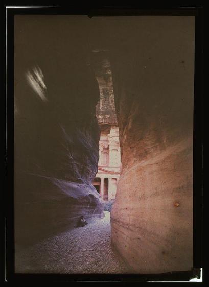 The image depicts a striking view of an ancient rock-cut architectural site, likely part of Petra in Jordan. The perspective is framed by towering sandstone cliffs that converge towards the center where an impressive, multileveled building emerges from within the rocks.
In this photograph's background lies what appears to be Nabataean architecture with its iconic facade and colonnade-style columns, indicative of ancient structures like the Monastery or Al-Khazneh. The foreground shows a narrow passage made up of smooth surfaces etched by erosion over time which suggests that the location could have been an ancient trade route.
The photograph is vintage in appearance, likely dating from between 1907 and 1946 as indicated by additional information provided about its source. It appears to be part of a collection housed at American Colony (Jerusalem), Photo Department, indicating it might belong to the Matson Collection or another historical archive.
The colors captured are warm earth tones with rich browns from the rock formations contrasted against the faded whites and grays of the Nabataean architecture in the background. The overall ambiance is one that transports viewers back through history, providing a glimpse into ancient civilization's architectural prowess.
