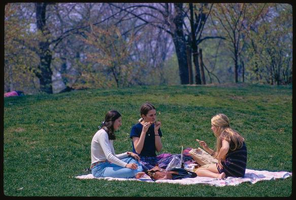 The image shows three women sitting on a blanket outside in the park during springtime. The woman in the middle wears blue and is eating while talking to her neighbors, one of whom has long brown hair with a ponytail secured by a scrunchie, wearing jeans and a white sweater over it. She looks at something off-camera. Her neighbor to the right appears blond and dressed in a striped sleeveless dress; she's holding some food that seems like a sandwich while looking down.

In front of them on the blanket are various items: another brown paper bag with bread or pastries, a cup possibly containing tea or coffee sitting beside what looks like a glass bottle. A plastic bag is also next to these objects along with other indistinct and blurry belongings.

Behind this group, trees can be seen that appear not fully in bloom but show the early signs of spring. The grass on which they are seated appears well-maintained, indicating it might be an urban park setting or a public area regularly kept for visitors' use.

In the background, another person seems to have their back turned and is possibly lying down; however, only partial visibility suggests this individual's presence in what looks like a relaxed scene.