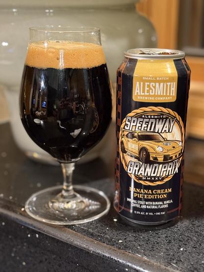 A snifter glass full of dark beer with a bubbly tan head. The black pint can has yellow accents and a yellow race car in its center